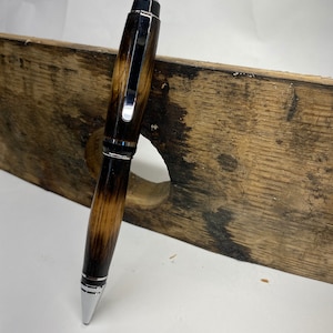 K&ouml;nnte beinhalten: Ein brauner und schwarzer Holzstift mit silbernen Akzenten. Der Stift liegt auf einem St&uuml;ck Holz.