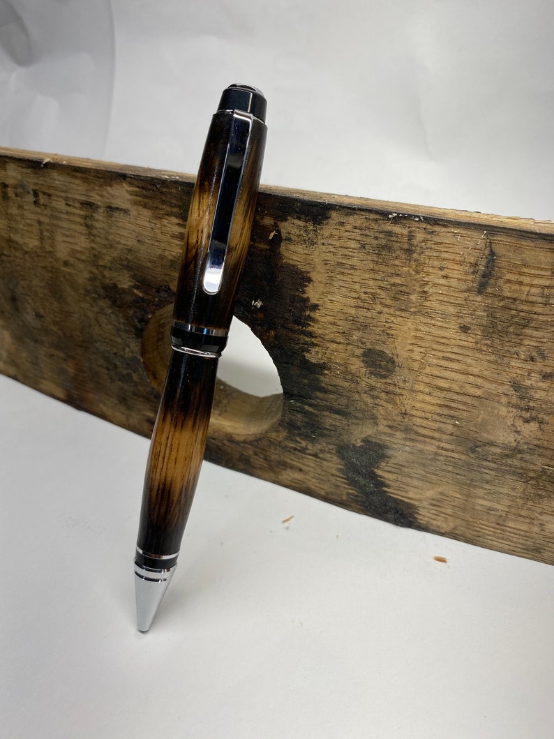 K&ouml;nnte beinhalten: Ein brauner und schwarzer Holzstift mit silbernen Akzenten. Der Stift liegt auf einem St&uuml;ck Holz.