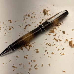 K&ouml;nnte beinhalten: Ein Holzstift mit silbernem Clip und schwarzen Akzenten. Der Stift liegt auf einer wei&szlig;en Oberfl&auml;che mit Holzsp&auml;nen, die um ihn herum verstreut sind.