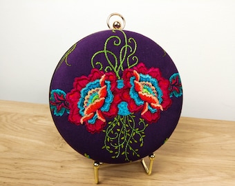 May include: A round, purple clutch purse with a gold clasp and floral embroidery. The embroidery features red, orange, pink, blue, and green flowers.