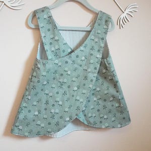 Könnte beinhalten: Ein wendbares Babykleid mit floralem Muster auf mintgrünem Hintergrund. Das Kleid hat ein Cross-Over-Design und hängt an einem hellblauen Kleiderbügel. Die Rückseite hat ein Streifenmuster.