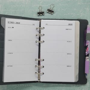 Könnte beinhalten: Ein weißer Planer mit schwarzem Einband und silbernen Ringen. Der Planer ist auf den Dezember/Januar-Seiten geöffnet. Die Seiten sind leer und haben ein gepunktetes Rastermuster. Der Planer hat eine Lasche mit der Aufschrift "WICHTIG" mit einem Herzsymbol. Der Planer hat auch eine Lasche mit der Aufschrift "JANUAR" mit den Wochentagen.