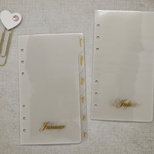 Może przedstawiać: Dwa przezroczyste, plastikowe separatory do planera ze złotymi literami dla miesięcy, w tym "Januar" i "Juli". Złoty spinacz do papieru z białym sercem i dżetem znajduje się w lewym górnym rogu. Separatory mają wstępnie dziurkowane otwory.