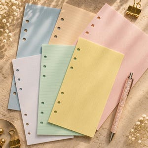 Könnte beinhalten: Eine Sammlung von pastellfarbenen Planer-Einlagen in Blau, Pfirsich, Rosa, Lavendel, Grün und Gelb. Die Seiten haben seitliche Lochungen und sind sowohl liniert als auch gepunktet. Ein glitzernder Stift und goldene Büroklammern sind ebenfalls enthalten.