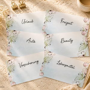 Könnte beinhalten: Sechs rechteckige weiße Karten mit hellblauem Aquarellverlauf und floralen Randverzierungen. Die Karten tragen die schwarze handschriftliche Beschriftung Urlaub, Freizeit, Auto, Beauty, Versicherung und Lebensmittel, angeordnet in zwei Spalten.