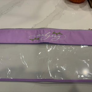 Può includere: Una custodia rettangolare color lavanda con un pannello frontale in plastica trasparente e una chiusura a zip. La sezione superiore in tessuto lavanda presenta la parola mahjong scritta in corsivo bianco, con piccole foglie verdi.