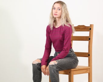 May include: A long-sleeved, striped top in shades of magenta and black. The top is paired with distressed gray jeans. The person is seated on a wooden chair with a light brown finish.
