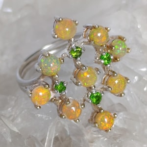 Könnte beinhalten: Ein silberner Ring mit einem Cluster-Design, besetzt mit mehreren runden, gelb-orangen Opalen und kleinen, runden grünen Edelsteinen. Die Opale weisen eine schillernde Qualität auf, und der Ring ist vor einem weißen Hintergrund platziert.