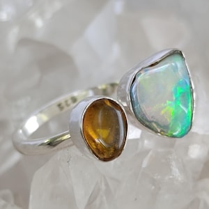 Könnte beinhalten: Ein Silberring mit zwei Edelsteinen. Einer ist ein großer, schillernder Opal mit grünen und blauen Reflexen. Der andere ist ein kleinerer, bernsteinfarbener Stein. Der Ring hat ein modernes, asymmetrisches Design.