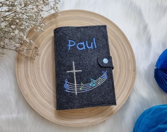 May include: A dark gray felt book cover with the name "Paul" embroidered in blue. A white cross and musical notes are embroidered below. The cover has a blue button closure. The cover is on a wooden plate.