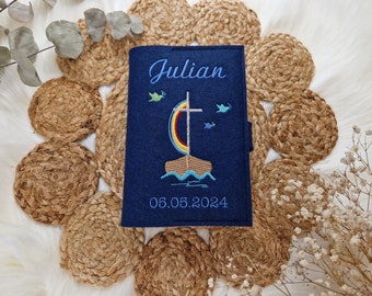 May include: A navy blue felt book cover with the name "Julian" embroidered in light blue script. The cover features a colorful embroidered boat with a cross and the date 05.05.2024. The cover is displayed on a woven mat.