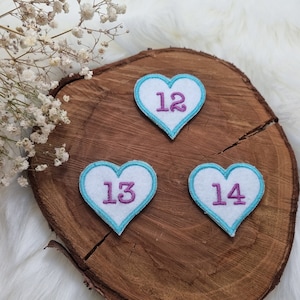 Könnte beinhalten: Drei weiße Filz-Herz-Patches mit blauen Umrandungen und violetten Zahlen. Die Zahlen sind 12, 13 und 14. Die Patches befinden sich auf einer Holzoberfläche.