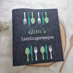 Könnte beinhalten: Ein grauer Filz-Rezeptordner mit dem Text "Gitti's Lieblingsrezepte" und einem Design aus grünen und weißen Gabeln, Messern und Löffeln.