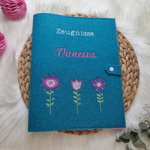 Könnte beinhalten: Ein blauer Filzordner mit einem rosa Knopfverschluss. Der Ordner ist mit Blumen bestickt und dem Text "Zeugnisse Vanessa" in weißer und rosa Farbe.