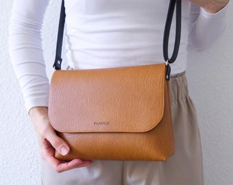 May include: A tan crossbody bag with a textured finish and a fold-over flap. The bag features the word PAMPLUS printed in black on the front flap and includes a thin black shoulder strap attached to the sides with silver-toned metal hardware.