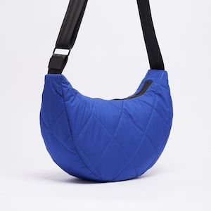 Op de afbeelding: Een blauwe gewatteerde schoudertas met een verstelbare zwarte schouderband. De tas heeft een ritssluiting en een halvemaanvorm.