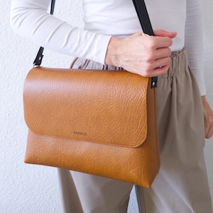 Könnte beinhalten: Eine hellbraune Umhängetasche mit strukturierter Oberfläche und schwarzem Schulterriemen. Die Tasche hat einen Klappverschluss und das Wort "PAMPUS" ist auf der Vorderseite sichtbar.