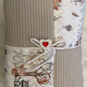 May include: A beige waffle weave baby blanket with a white fabric panel featuring a woodland animal print. The blanket is tied with a white ribbon and a red heart-shaped tag.