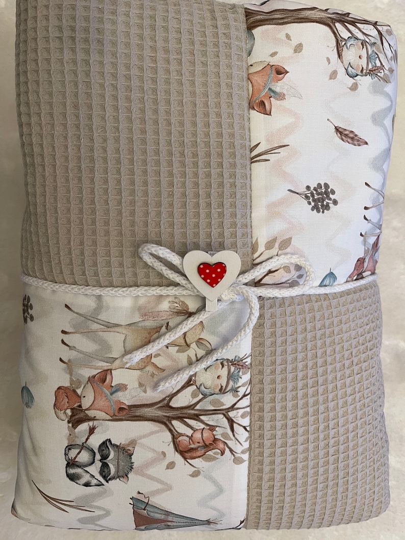 May include: A beige waffle weave baby blanket with a white fabric panel featuring a woodland animal print. The blanket is tied with a white ribbon and a red heart-shaped tag.