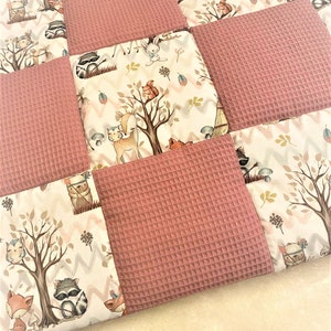 Könnte beinhalten: Eine rosa-weiße Patchwork-Babydecke mit Waldtier-Motiv. Die Decke zeigt verschiedene Tiere, darunter Rehe, Füchse, Waschbären und Eulen. Die Decke ist aus weichem, bequemem Stoff und perfekt zum Kuscheln.