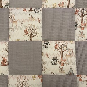 Op de afbeelding: Een patchworkdeken met een bosmotief. De deken is gemaakt van verschillende stoffen, waaronder een grijs wafelstof en een cr&egrave;mekleurige stof met een bosdierprint. De deken heeft verschillende bosdieren, waaronder herten, wasberen, vossen en uilen.