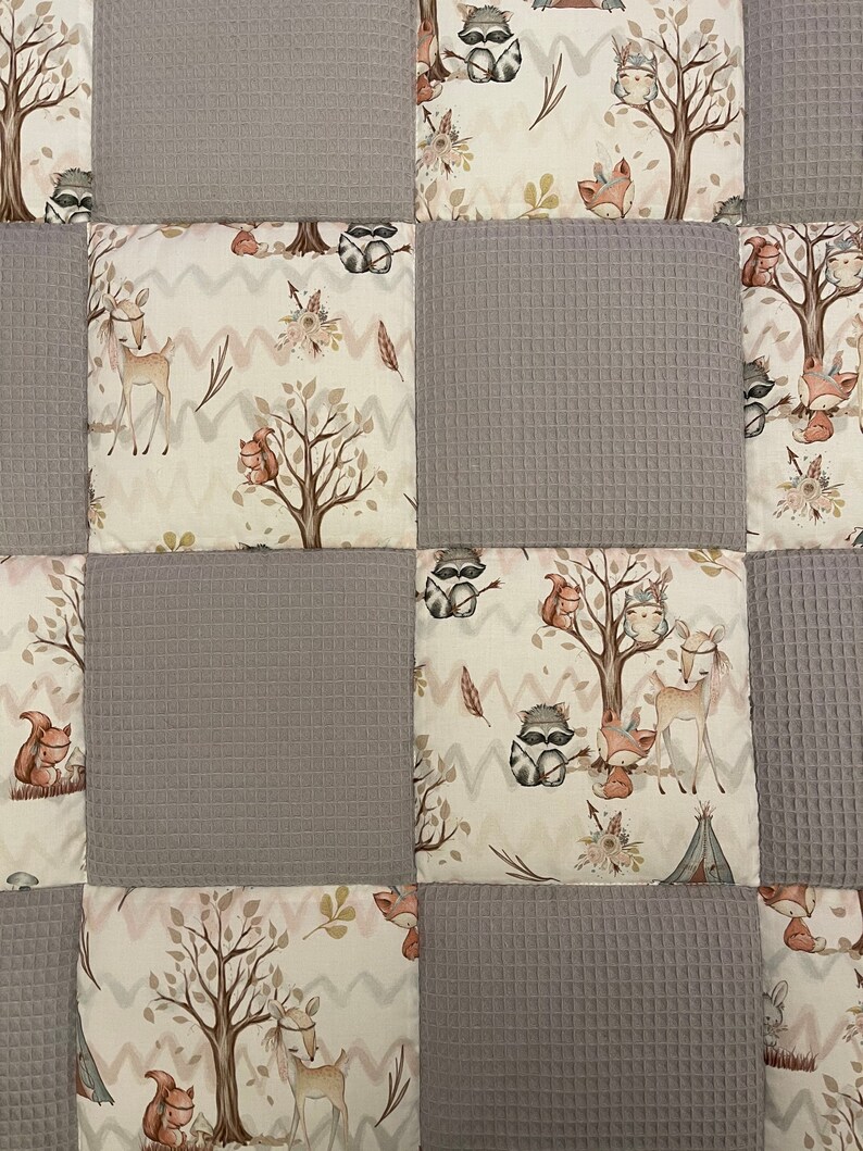 Op de afbeelding: Een patchworkdeken met een bosmotief. De deken is gemaakt van verschillende stoffen, waaronder een grijs wafelstof en een cr&egrave;mekleurige stof met een bosdierprint. De deken heeft verschillende bosdieren, waaronder herten, wasberen, vossen en uilen.