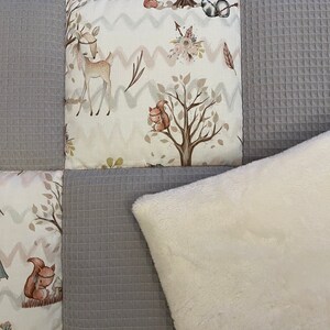 Op de afbeelding: Een grijs en wit patchwork-deken met een wit nep bont kussen. Het deken heeft een bosdieren thema met een hert, eekhoorn en wasbeer.