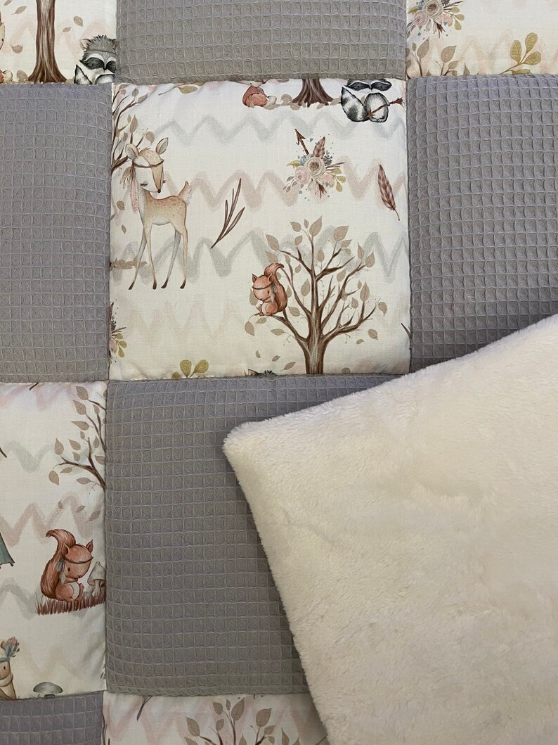 Op de afbeelding: Een grijs en wit patchwork-deken met een wit nep bont kussen. Het deken heeft een bosdieren thema met een hert, eekhoorn en wasbeer.