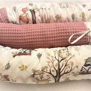 Könnte beinhalten: Ein rosa-weißer Babybett-Stoßfänger mit einem Waldtier-Druck. Der Stoßfänger ist aus weichem, gestepptem Stoff gefertigt und zeigt eine Vielzahl von Tieren, darunter Eulen, Waschbären und Rehe. Der Stoßfänger ist mit einem weißen Band zusammengebunden.
