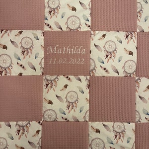 Könnte beinhalten: Eine rosa-weiße Babydecke mit Patchwork-Muster. Jedes Quadrat hat einen weißen Hintergrund mit einem Traumfänger- und Federmuster. Das mittlere Quadrat ist rosa und hat den Namen "Mathilda" und das Datum "11.02.2022" aufgestickt.
