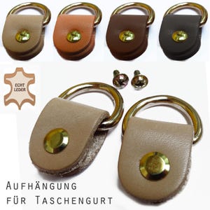 Könnte beinhalten: Leder-Taschenriemen-Befestigungen in verschiedenen Farben: Beige, Orange, Braun und Dunkelbraun. Jede Befestigung hat einen goldfarbenen Metallring und eine passende goldfarbene Niete. Der Text "ECHT LEDER" und "AUFHÄNGUNG FÜR TASCHENGURT" ist ebenfalls enthalten.