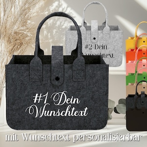 Könnte beinhalten: Eine dunkelgraue Filztasche mit zwei Henkeln und einem mittigen Druckknopfverschluss. Auf der Vorderseite steht der weiße Text #1 Dein Wunschtext. Dahinter ist eine hellgraue Filztasche mit dem Text #2 Dein Wunschtext sowie ein Stapel bunter Taschen.