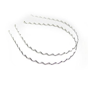 Könnte beinhalten: Zwei silberne, gewellte Haarbänder, jedes mit einem zarten, leicht gekrümmten Design. Die Haarbänder sind in einem lockeren Kreis angeordnet, wobei sich ein Haarbänder über das andere legt.