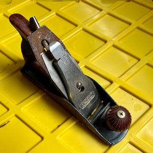 May include: A vintage wood plane with a metal blade and a brown wooden handle. The plane is labeled "CORSAIR" and has a metal knob on the side.