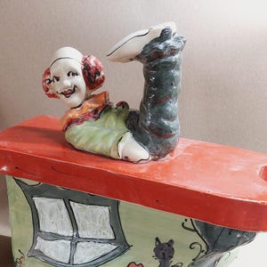 Könnte beinhalten: Skurrile Keramikfigur mit einem lächelnden Clown auf einem Miniaturhaus. Das Haus hat ein rotes Dach, grüne Wände und ein Fenster. Auf der Seite ist eine schwarze Katze bemalt, mit Blumen- und Grasdetails.