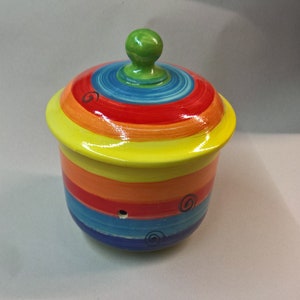 May include: A ceramic jar with a lid, painted with horizontal bands of bright colors: yellow, orange, red, blue, and green. The lid has a green knob. The jar has a small hole and decorative swirls.