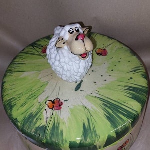 Peut inclure: Une boîte ronde en céramique avec un motif peint vert et blanc. Le couvercle présente un mouton en céramique blanc avec un nez rouge et des yeux noirs.