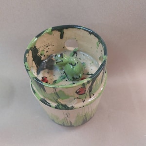 Puede incluir: Un pequeño macetero de cerámica con una figurita de rana verde en la parte superior. El macetero está pintado con pintura verde, blanca y negra salpicada.