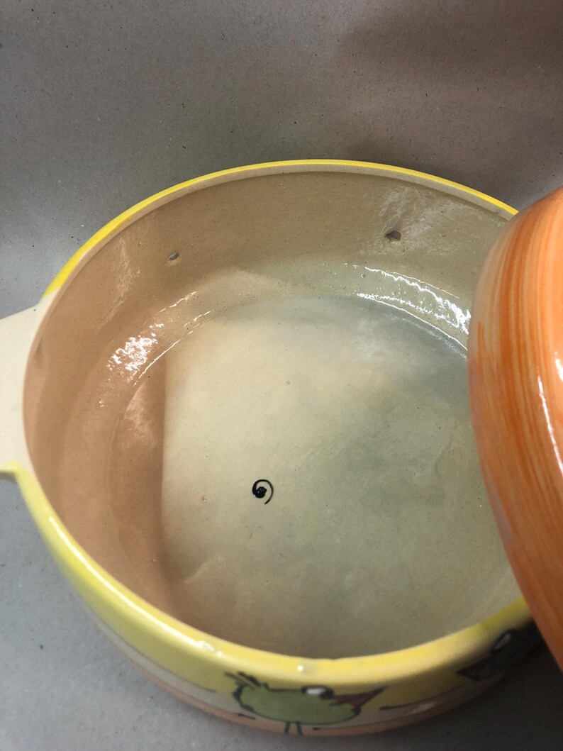 May include: A yellow ceramic casserole dish with a lid. The dish has a brown interior and a colorful design of birds and frogs around the rim.