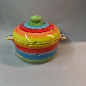 May include: A ceramic casserole dish with a lid, painted with concentric bands of rainbow colors. The dish has two small handles and a green knob on the lid. The design includes black swirl accents.