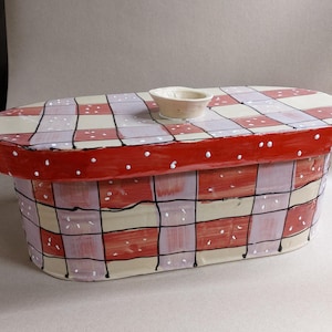May include: Hand-painted ceramic bread bin with a red and cream checkered pattern. The lid is red with white polka dots. A small, matching bowl sits on top. The bin is oval-shaped and has black lines outlining the squares.