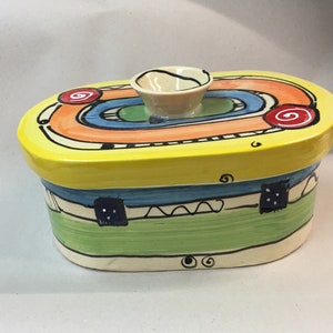 Puede incluir: Un recipiente de mantequilla ovalado de cerámica amarilla con un diseño colorido de naranja, azul, verde y rojo. La tapa tiene un pequeño pomo de cerámica blanco.