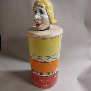 Peut inclure: Pot en céramique décoratif avec un couvercle en forme de tête de femme. Le pot est peint avec des bandes horizontales de jaune, d'orange et de rouge, avec des pois blancs sur la section rouge. Le couvercle est de couleur bois clair.