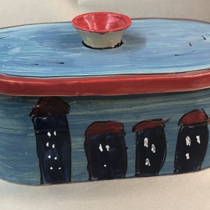 Könnte beinhalten: Ein blauer und roter Keramikbehälter mit Deckel. Der Behälter hat ein gemaltes Design von vier Gebäuden mit roten Dächern. Der Behälter ist oval.