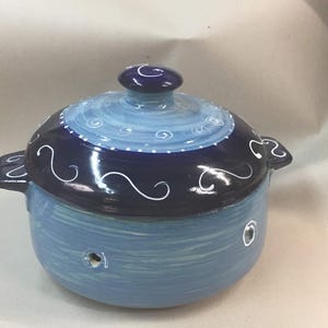 Peut inclure: Une marmite en céramique bleue avec un couvercle. Le pot est peint dans des tons de bleu, avec une base bleu clair et un couvercle bleu foncé. Des motifs tourbillonnants blancs ornent le couvercle. La marmite a deux petites poignées.