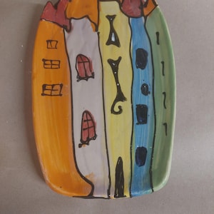 Könnte beinhalten: Handbemalte Keramikplatte mit einem skurrilen Stadtlandschaftsdesign. Die Platte zeigt farbenfrohe Gebäude in Orange-, Weiß-, Gelb-, Blau- und Grüntönen mit roten und orangen Dächern. Die Platte ist oval geformt.