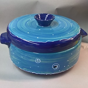 May include: A hand-painted ceramic casserole dish with a lid. The dish is primarily turquoise and blue, with white accents. The lid has a small handle and a wavy white border. The dish has two handles on the sides.