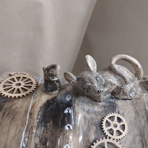 May include: A ceramic sculpture of two mice and wooden cogs. The larger mouse is grey and brown, with its ears pricked. The smaller mouse is black and grey. The cogs are made of light-coloured wood. The sculpture has a rustic, handmade appearance.