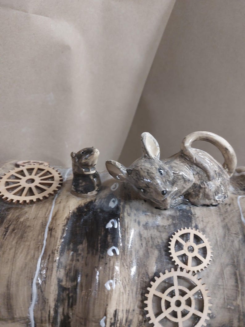 May include: A ceramic sculpture of two mice and wooden cogs. The larger mouse is grey and brown, with its ears pricked. The smaller mouse is black and grey. The cogs are made of light-coloured wood. The sculpture has a rustic, handmade appearance.