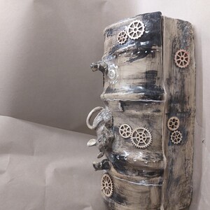 May include: A cylindrical ceramic sculpture in a steampunk style. It has a textured, aged appearance with black and beige tones. Wooden gear accents and a small animal sculpture are incorporated into the design.